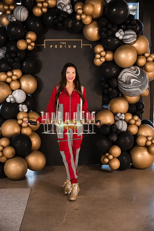 Woman smiling and holding a tray of champagne glasses in front of a photo-op balloon wall